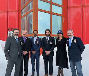 Six people in suits, with one lady wearing a long black dress, stand side-by-side in front of a red steel building. The people all have red poppy badges on their clothing and some have service medals of various colours. There is a full cover of snow on the ground, also built up against the side of the building.