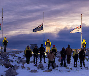 Three flag poles occupy the main part of the photo, with flags at half mast. There is a group of 13 people standing in front of the flags and facing the camera. The flags are all Australian, with the right hand one being white for the Navy, the centre one is the dark blue Australian Flag, and the left hand flag is light blue for the Airforce. The flag poles are mounted on a snow covered rocky hill and the sky is partly cloudy.