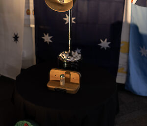 The cenotaph for our inside service. It is an Army slouch hat, resting atop an ice-pick. The pick is inserted into a black plant pot filled with white stones, and is sitting on a low round table with a black table cloth covering it. There is a small glass container with sand from the beach at Gallipoli sitting in front of the plant pot. On the ground in front of the table are two green circular wreaths, with red poppies on them. The background is the Australian Flag, hanging from the stairs that are out of view above.