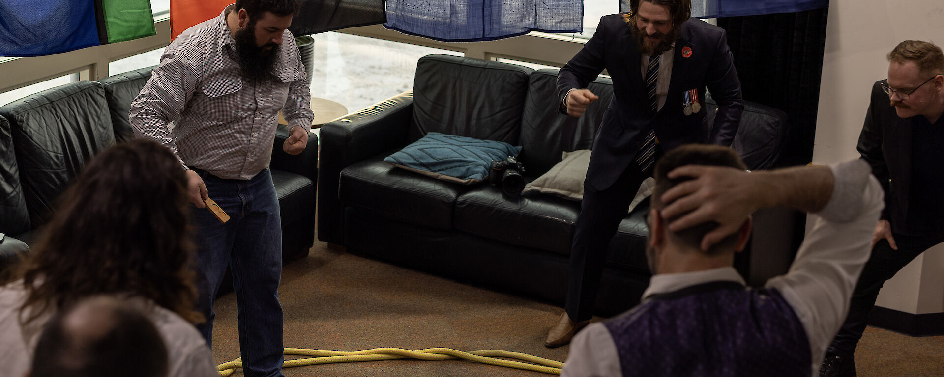 Five men are standing around a circle of yellow rope laid out on the brown carpet. One man has a small wooden paddle in his hand and there is a coin in the centre of the shot, on the ground. The men are in excited, animated poses, focused on the coin.