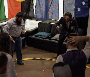 Five men are standing around a circle of yellow rope laid out on the brown carpet. One man has a small wooden paddle in his hand and there is a coin in the centre of the shot, on the ground. The men are in excited, animated poses, focused on the coin.