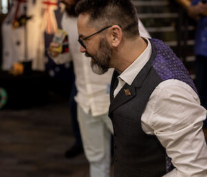 A man in a white shirt and dark grey vest holds a fake money note above his head, hoping for the coins to land on heads. He is staring intently to the left of the photo, where the two-up ring is located out of frame. In the blurred background stands another man also focused on the out of frame action.