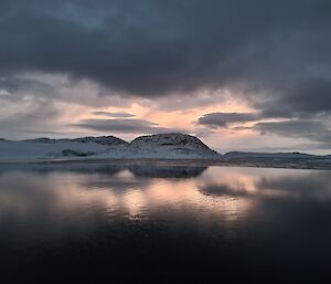 The lower half of this photo is calm glassy dark grey water, reflecting the cloud filled sky above. Across the centre of the photo is some low rocky snow covered hills. The sunlight filtering through the clouds gives the scene a light purple-grey colour.