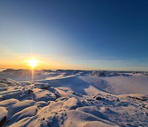 Snow covered rocky hills stretch from the bottom of frame to the centre. The bright yellow-orange sun sits just above the horizon, with thin yellow beams extending out from its centre into the clear blue sky.