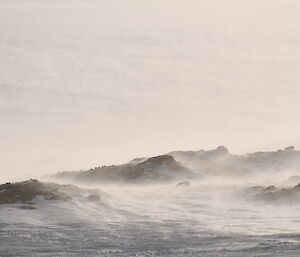 Low jagged black rocks stretch across the frame from left to right. They protrude shallowly from the snow filling the rest of the photo. The wind is sweeping snow across the rocks from the left.