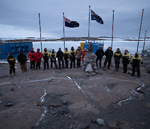 Sixteen people stand in front of the Australian and New Zealand flags with a frozen harbour in the background