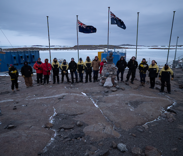 Sixteen people stand in front of the Australian and New Zealand flags with a frozen harbour in the background