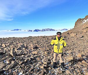 A man is standing on rocky terrain with ice and rocky mountain range in the distance