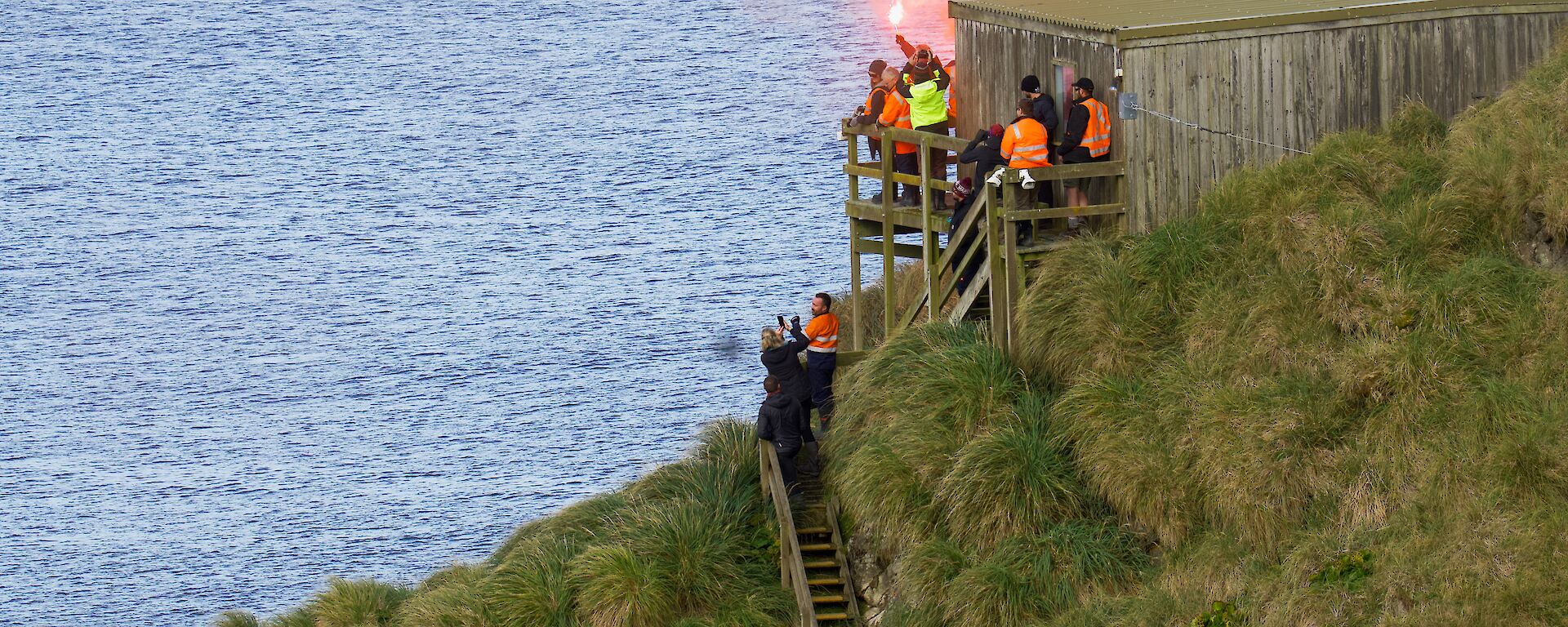 A group of people near a wooden hut by the ocean let off orange flares to farewell a ship