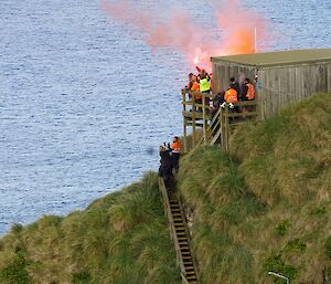 A group of people near a wooden hut by the ocean let off orange flares to farewell a ship