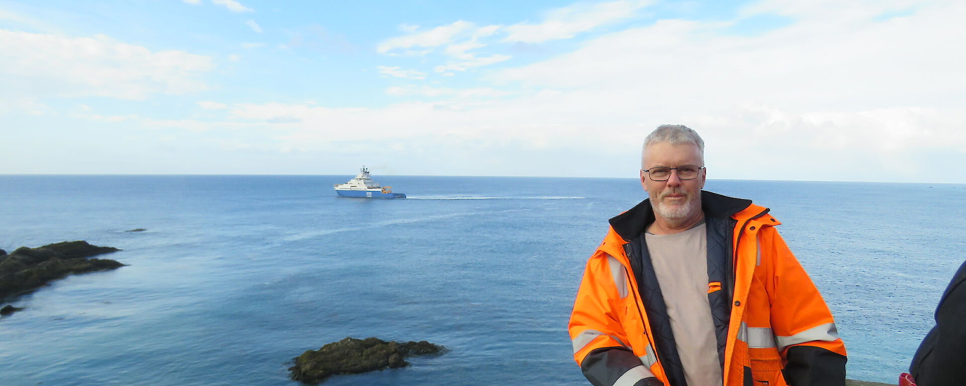 A man in hi vis stands in front of a blue seascape with a ship in the distance