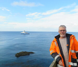 A man in hi vis stands in front of a blue seascape with a ship in the distance