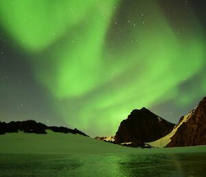 An green aurora dances in the sky above a frozen lake with rocky peaks surrounding