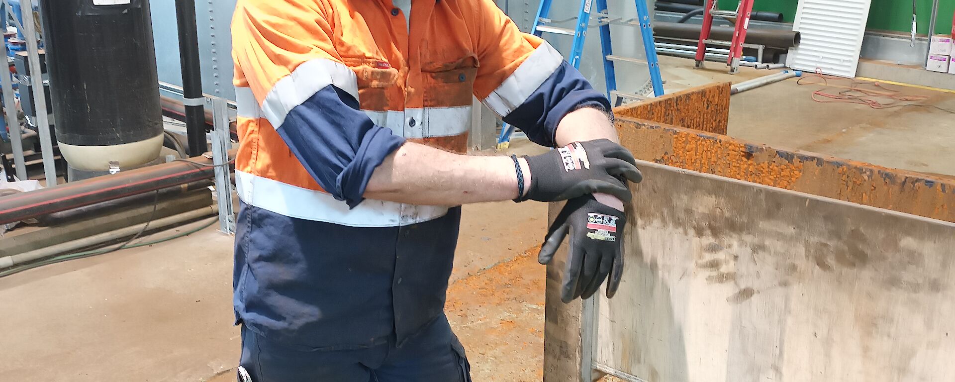 A man in orange hi-vis workwear is leaning on a white steel workbench. He is wearing a black baseball cap and glasses. In the background there is a large silver steel water tank and pipes stretching across the photo.