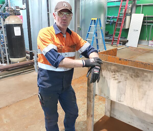 A man in orange hi-vis workwear is leaning on a white steel workbench. He is wearing a black baseball cap and glasses. In the background there is a large silver steel water tank and pipes stretching across the photo.