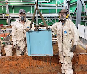 In the centre of the photo is a light blue square steel metal plate, hanging from a crane hook. Either side of the plate is two men in disposable white overalls, respirators and safety goggles. They are quite dirty. They are standing in the remains of the water tank, which is a rust covered concrete block. The green walls of the shed are in frame.
