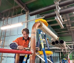 A man in orange hi-vis is standing on a ladder, facing the camera. He is working on some large copper pipes with a wrench. In the background is a very large light blue water tank made of square metal panels bolted together.