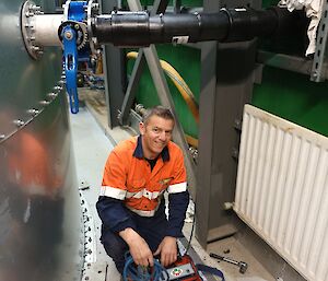 A man in orange hi-vis workwear is kneeling down and looking up at the camera smiling. He is holding some electrical test equipment. To the left is the side of a large silver steel water tank. To the right is the framework of the shed he is working in, and the green shed cladding is also visible.