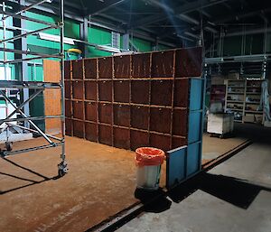 In the centre of this photo is a large wall made of rusted square metal plates. A rust covered concrete floor extends either side of this wall. In the background is the green wall of the shed.