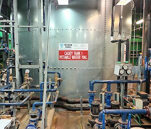 The rear of this photo is filled with a very large 70000 litre bare steel cylindrical water tank. The foreground is filled with pipework and small pumps. Overhead there are two long florescent lights.