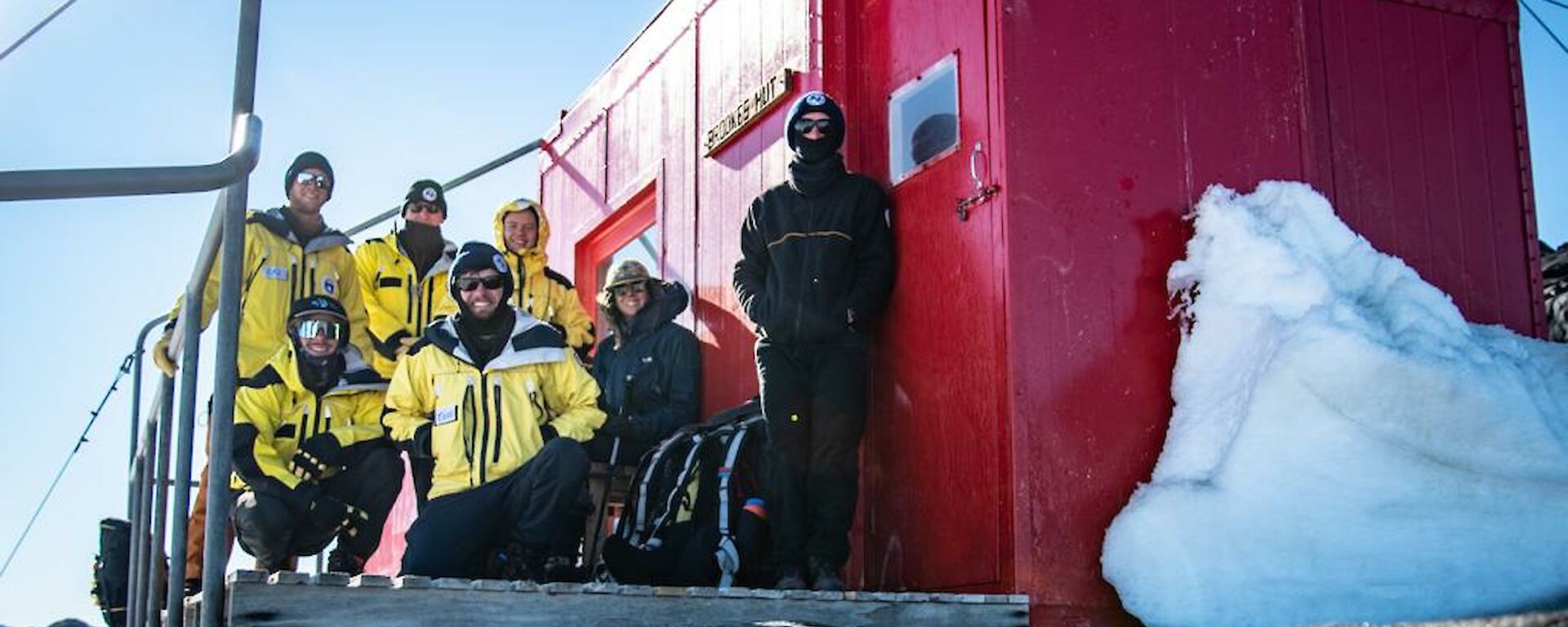 A group of smiling people in yellow cold weather clothing stand on the deck of a red field hut.