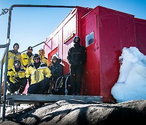 A group of smiling people in yellow cold weather clothing stand on the deck of a red field hut.