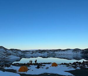Two yellow tents and a group of people in the snow next to a lake surrounded by snow-covered hills