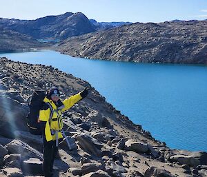 A smiling man wearing yellow cold weather clothing giving the peace sign and standing next to a long blue lake surrounded by rocky hills.