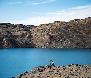 Two hikers standing by a blue lake surrounded by rocky hills.