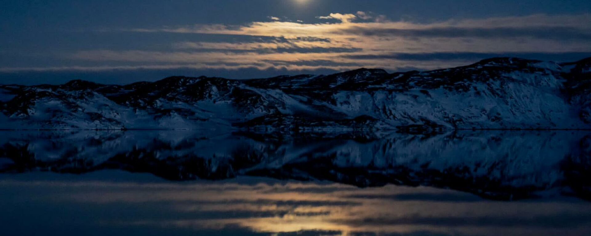The moon rising over snow-covered hills and reflecting off the still waters of a lake.