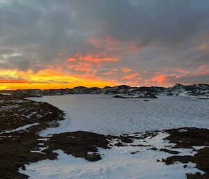 A frozen fjord surrounded by snow covered hills with the sun setting in the clouds in the background.
