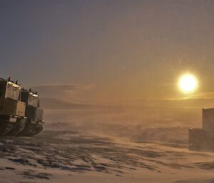 Snow is being blown across a icy landscape with the sun setting in the distance and two tracked vehicles on the left of frame