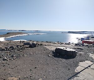 A harbour with blue water sits under a summer's sky with a rocky landscape in the foreground