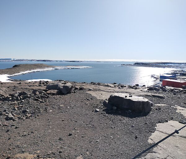 A harbour with blue water sits under a summer's sky with a rocky landscape in the foreground