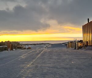 On the right of this wide framed photo is a large orange steel shed, with a yellow sunrise reflecting on it. On the left of the scene is a cylindrical fuel tank and some shipping containers. The centre of the photo is a wide snow covered road disappearing into the distance. The sky is cloudy but there is a horizontal band of yellow-orange light from the sunrise stretching across the image.