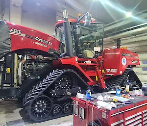 This photo is taken inside the mechanical workshop and the frame is almost completely filled with a very large red tractor. The tractor has tracks instead of wheels. In the foreground is a large red toolbox.