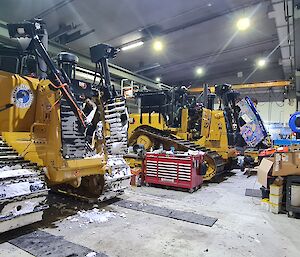 This photo is inside the mechanical workshop shed and shows two very big yellow bull dozers, with no blades, waiting in adjacent parking bays for a service. They have steel tracks that are covered in snow, and there is snow on the concrete floor of the workshop.