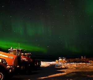 The lower left of the photo shows three large red tractors in a row. It is night time and the sky is full of stars, and vertical streaks of green from an aurora. In the distant right of the photo there are a couple of large yellow bull dozers.