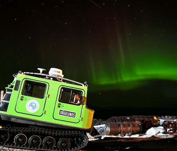 A square green vehicle with tracks like a tank is filling the bottom left of the photo. It is night time and the sky is filled with stars and also vertical streaks of green and red, generated by particles from the sun hitting our atmosphere. In the right foreground are a couple of large steel loader buckets off some heavy machinery.