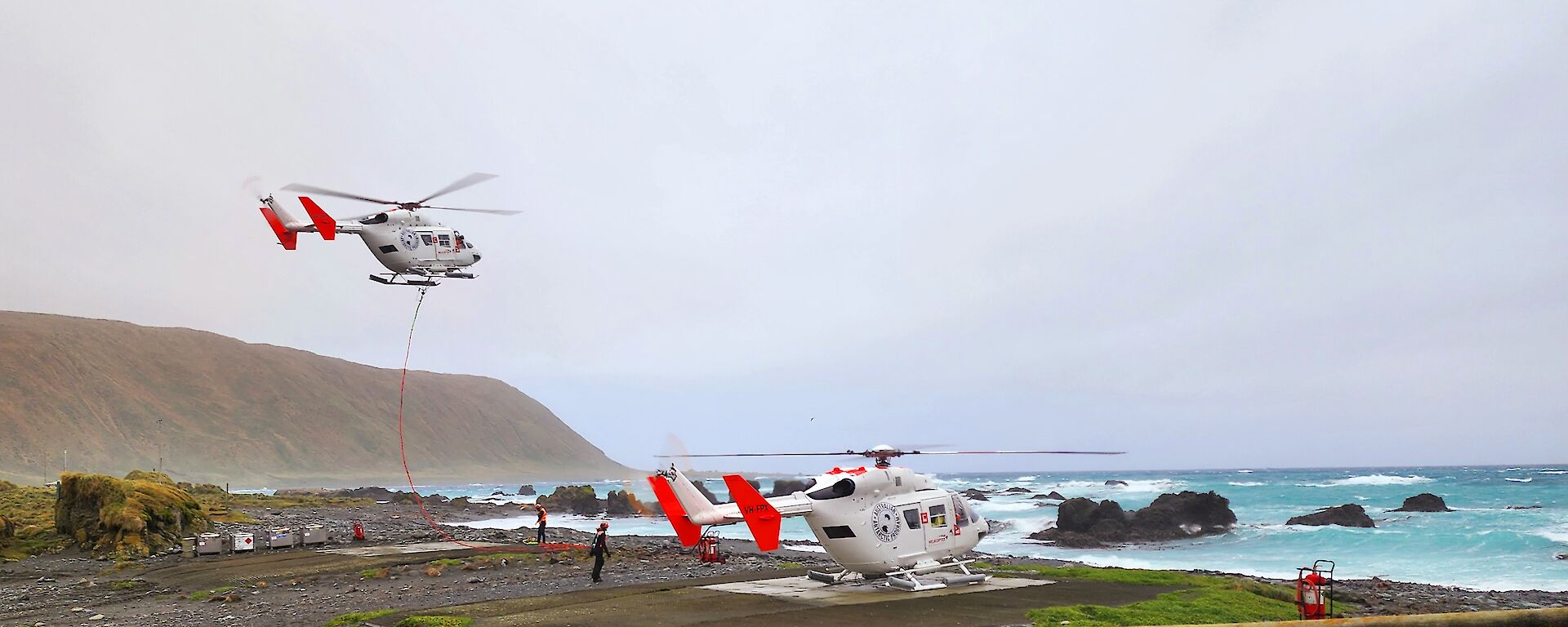 Two helicopters are close to the shoreline of a rugged island, one on the ground and one flying above