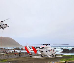 Two helicopters are close to the shoreline of a rugged island, one on the ground and one flying above