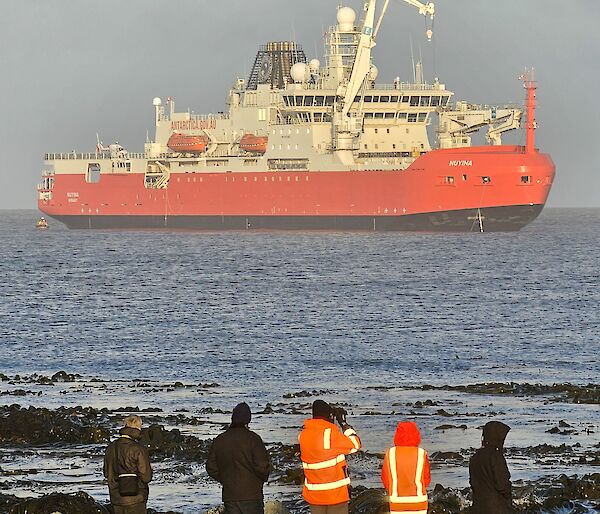 A group of people stand on the shore in front of a large red and white ship