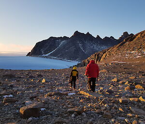 Three people are walking across a rocky landscape with mountain peaks and ice in the distance