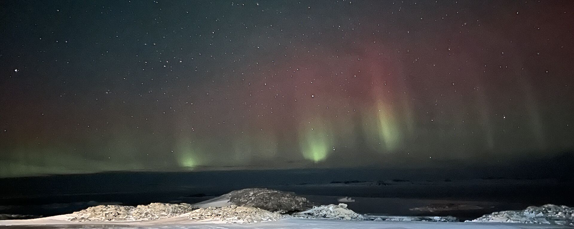The foreground of this photo is a lightly snow covered rocky piece of ground. It is night time and the sky in the background is full of stars. There are tall vertical beams of light that are green at the base, transitioning into red at the tops. They vary in height, with the tallest filling the middle third of the picture.