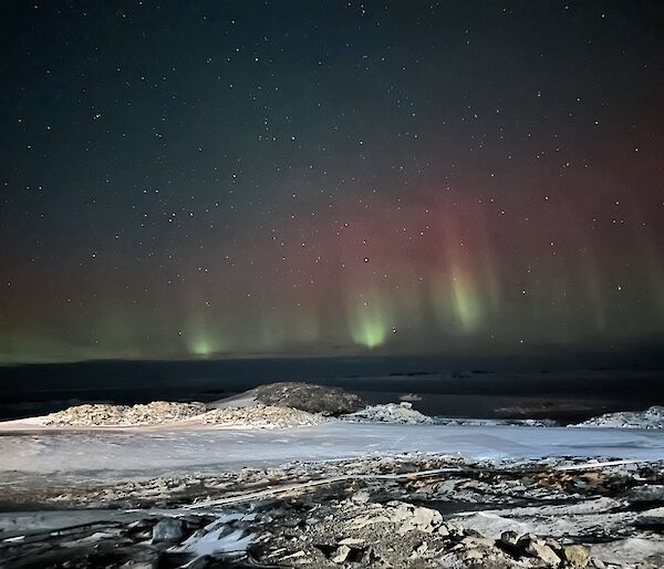 The foreground of this photo is a lightly snow covered rocky piece of ground. It is night time and the sky in the background is full of stars. There are tall vertical beams of light that are green at the base, transitioning into red at the tops. They vary in height, with the tallest filling the middle third of the picture.
