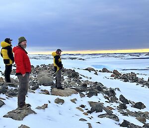 The lower two thirds of the photo are filled with sloping rocky ground, partially covered in snow. The top third is an overcast sky, with a thin ribbon of clear light yellow sky separating the two. There are three men to the left of the picture, all looking towards the right hand side of the picture. The men are dressed warmly and all have their hands in their pockets.