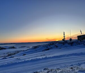 This photo is divided in two horizontally. The top half is clear sky, blue at the top and transitioning to orange lower down, where the sun is just peaking over the horizon. The lower half of the photo is of snow covered ground of Casey Station, and low rocky hills. There is a large green metal building to the right of the frame, and a flag flying adjacent to it.