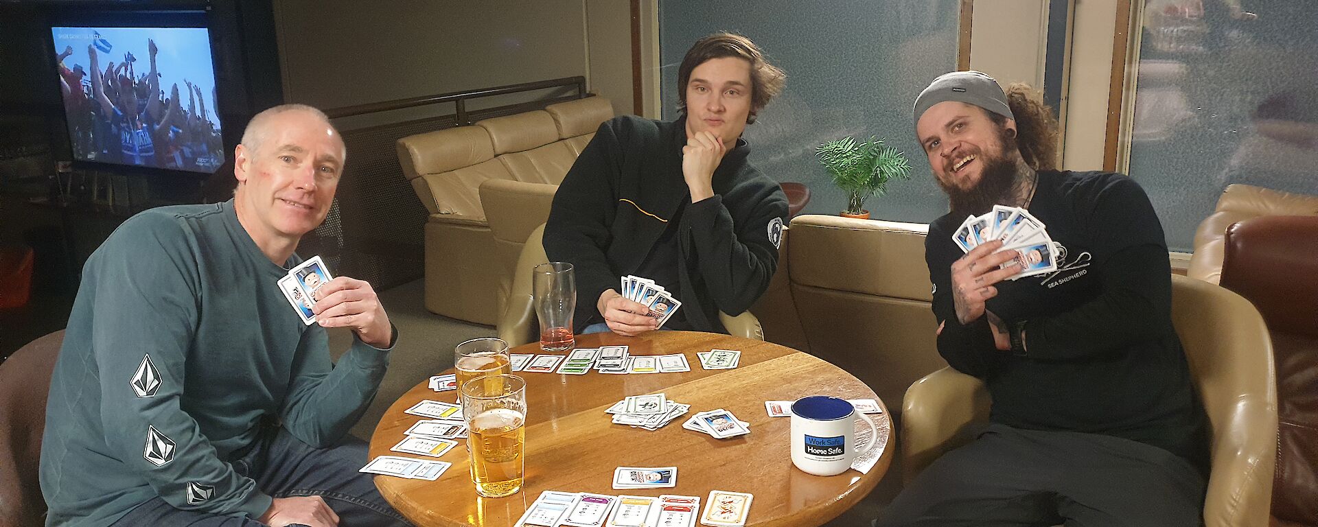 Three smiling men playing cards sit around a round bar table.