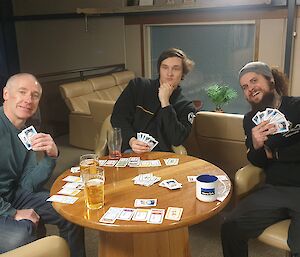 Three smiling men playing cards sit around a round bar table.