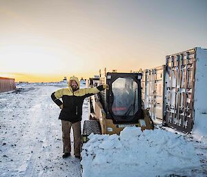 A smiling man wearing yellow snow clothes stands next to a yellow digger in the snow.